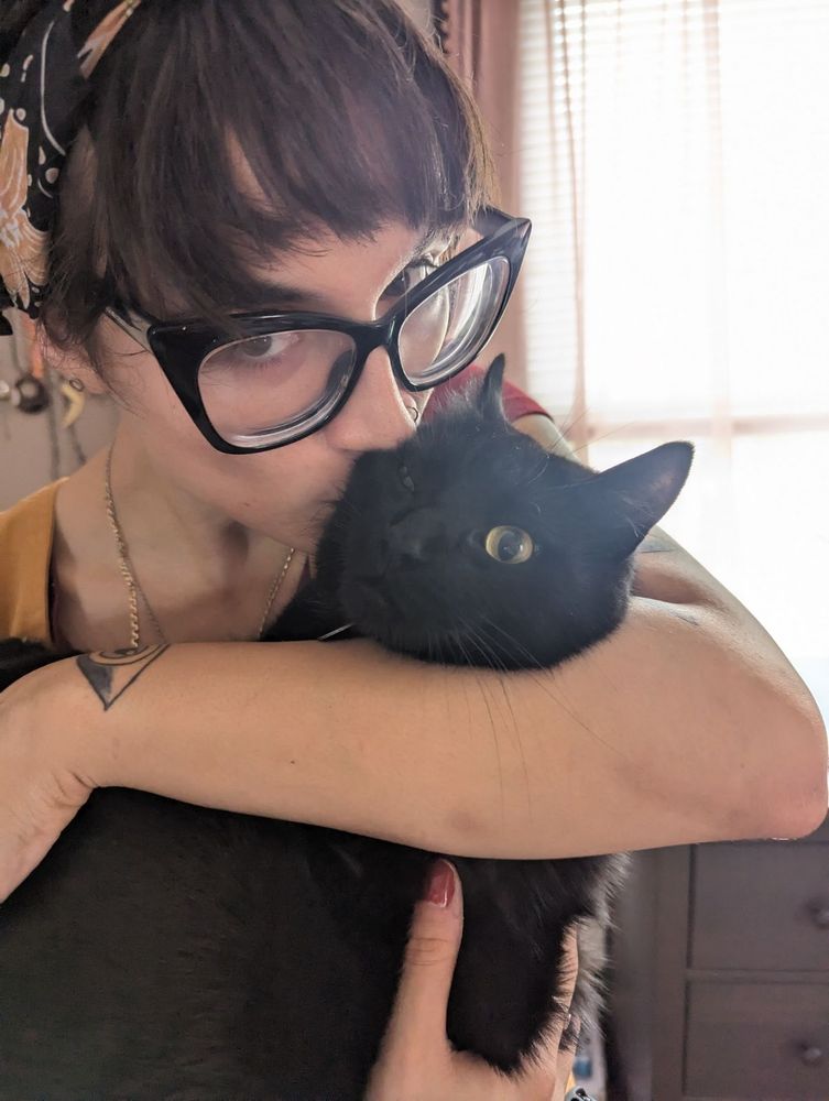 Ari in brown overalls and a floral bandana kissing a balck cat on the cheek