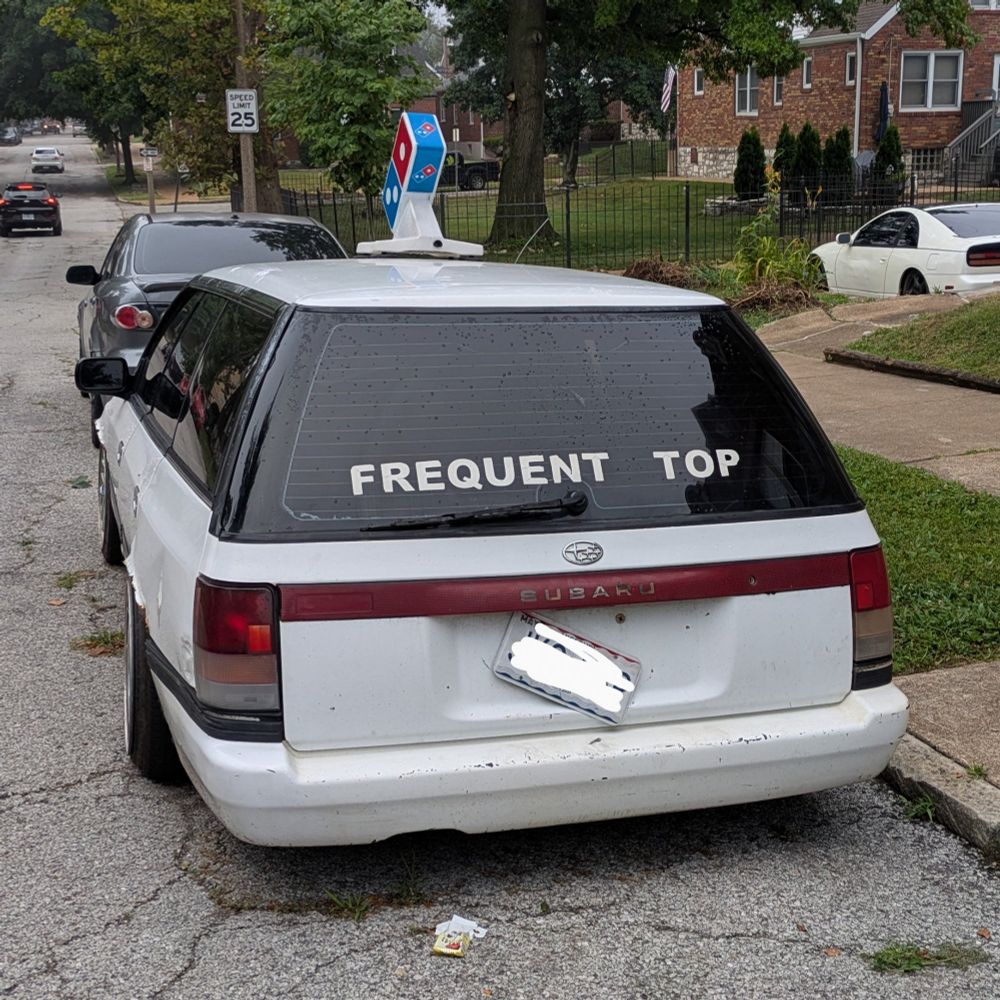 Photo of a white Subaru with "FREQUENT TOP" across the back window. There is a Domino's pizza light on the roof. It is morning.