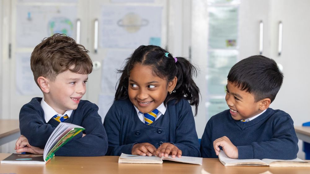 Three children in their school uniforms, smiling as they read together. 