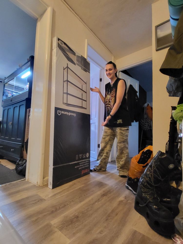 A photo of me in a hallway stood next to a large flat packed box with a rack shelving imaged on the front. The box is upside down.