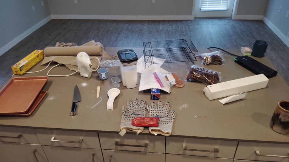 Messy kitchen island with an empty apartment living room in the background.
Cookie sheets, knife, cooling racks, few spots of spilled spices & flour, sugar container, hand mixer, even a pile of left over shelf liner all littering the island.