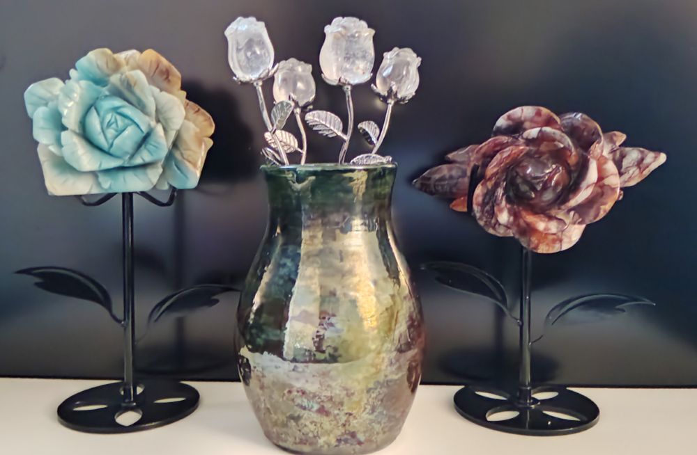 A raku vase (with some iridescent traits - reflecting multiple colors) holding four small, clear crystal roses with silver stems & petals. On the left is a larger blueish crystal rose on a stand and on the right is a reddish (with purple) crystal rose, also on a stand.