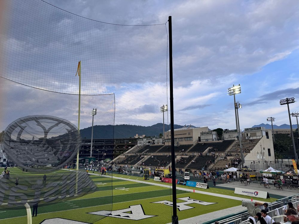 Photo of the home sideline of Hawaii’s home football stadium taken from end zone stands. It’s an hour before the game so not many people are here yet. A ridge line is visible above the stadium