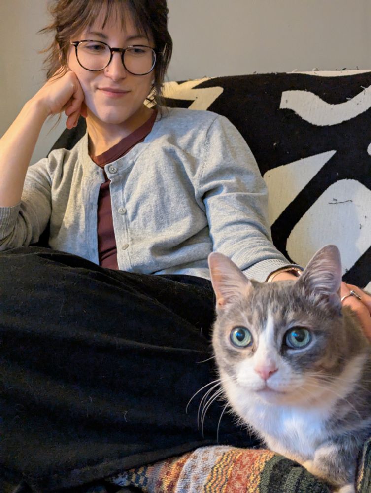 A photo of me curled up on a chair, smiling and looking down at a little gray and white cat with huge green eyes who is laying on my feet even though I’m a scary stranger.