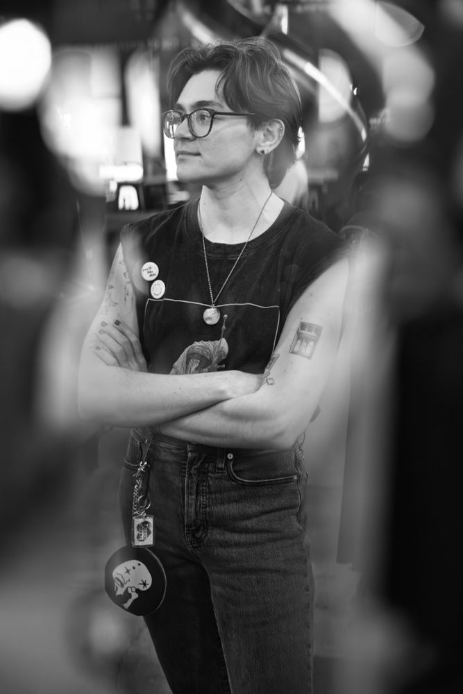 This is a stylish black and white photograph of a person standing with their arms crossed, looking off to the side with a thoughtful expression.

The person has dark, medium-length hair and is wearing glasses. Their outfit consists of a dark, sleeveless t-shirt (a muscle tee) adorned with several small pins, and dark-wash jeans. Tattoos are visible on both of their upper arms.

They are accessorized with a chain necklace holding a circular pendant and a distinctive round, dark pouch with a white graphic design hanging from their belt loop.

The photograph is composed with blurred elements in the foreground, which creates a sense of depth and frames the subject in a candid, observational style. The black and white format enhances the focus on texture and light, contributing to the cool, contemplative mood of the image.