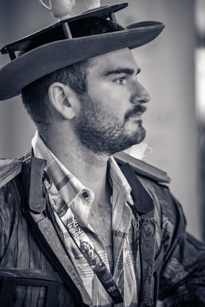 This is a striking black and white profile portrait of a man cosplaying as Able the Table from the game "Date Everything."

The man has a dark beard and mustache and is looking thoughtfully off to the right. He is wearing his distinctive wide-brimmed hat, which is uniquely adorned with a small platform holding white teacups.

His costume consists of a jacket with a prominent wood-grain texture worn over a plaid button-down shirt. The black and white format of the photograph emphasizes the rich textures of his attire and beard, lending a classic and dramatic quality to the image. The tight, profile composition creates an artistic and contemplative portrait, focusing on the character he is portraying.