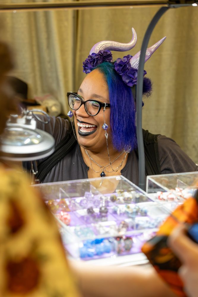 This is a lively and joyful photograph of a woman, who appears to be a vendor at a market or convention, sharing a laugh with someone.

She has a wonderfully expressive face, lit up with a wide, open-mouthed smile. Her unique and colorful style is prominent: she has blue and purple hair adorned with a headband of purple fabric flowers and light purple, iridescent horns. She wears black cat-eye glasses, which have decorative chains with dice-shaped charms, and her makeup includes bold black lipstick.

She is situated behind a table filled with clear display trays containing small, colorful dice that she is selling. An overhead lamp illuminates her and her wares. The back of a customer is visible in the foreground, indicating a warm and positive interaction. The image beautifully captures a moment of genuine happiness and connection in a creative, social setting.