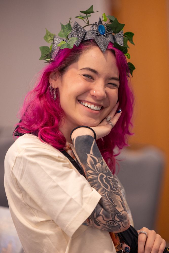 This is a cheerful and vibrant portrait of a person with a joyful expression.

The subject's most striking feature is their bright, magenta-pink hair, which is crowned with a unique, fantasy-inspired headpiece. The crown appears to be crafted from gray, stone-like segments interwoven with green ivy leaves, featuring a prominent blue gem set in the center.

They have a wide, radiant smile that crinkles their eyes, and they are looking directly at the camera. They have a small nose stud and are wearing multiple silver earrings and rings. Their head is tilted slightly, resting on their left hand.

A large black and grey tattoo sleeve is visible on their left forearm, featuring intricate designs and a solid black band around the wrist. They are dressed in a simple, cream-colored t-shirt, with a black bag strap crossing their chest.