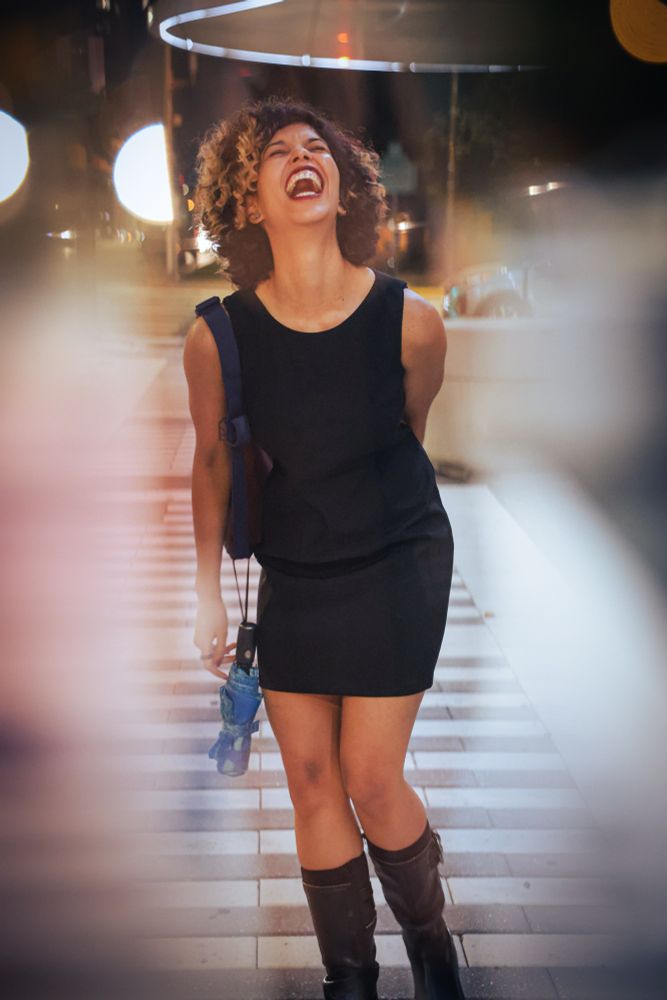 This is a candid and joyful photograph of a woman laughing wholeheartedly on a city street at night.

Her head is tilted back and her mouth is open in a genuine, exuberant laugh. She is wearing a sleeveless black dress and dark, knee-high boots. A blue strap rests on her shoulder, and she is holding a small, closed umbrella.

She is standing on a wide, modern crosswalk with bold white stripes. The background is softly blurred, showing the warm glow of city lights and creating a pleasant bokeh effect. The photo has an artistic quality, with a soft, hazy light leak or vignette framing the scene, which adds to the warmth and spontaneity of the moment.