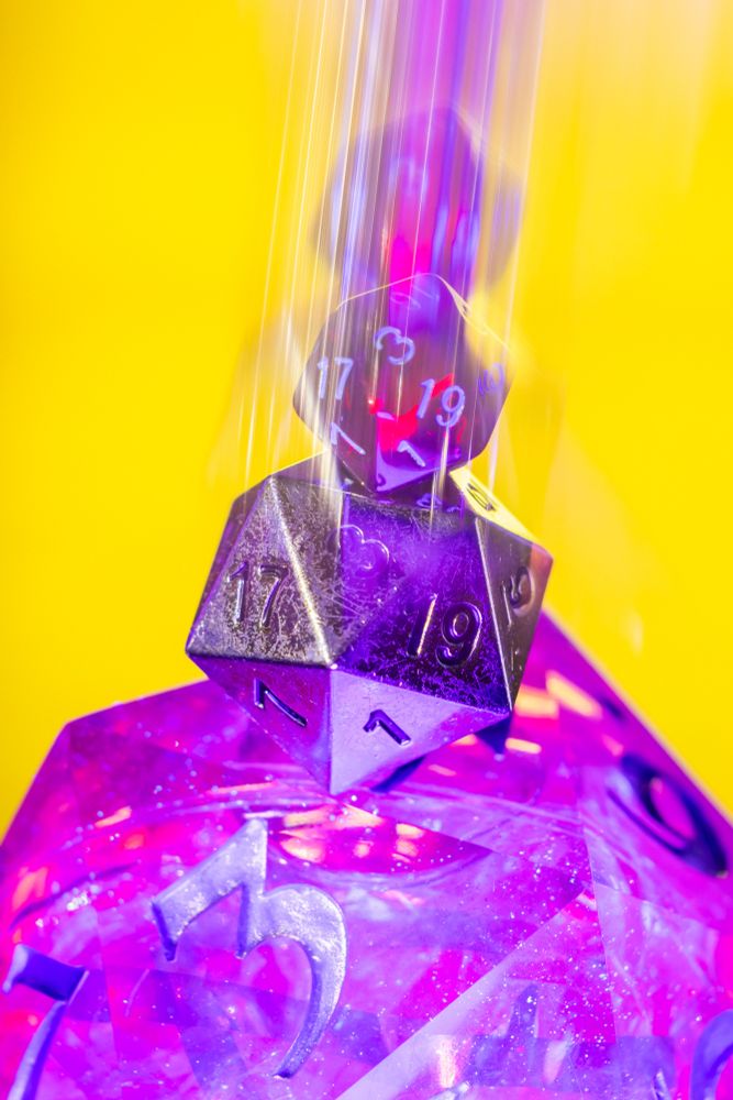 An artistic, close-up photograph of a tower of Dungeons & Dragons-style dice. At the bottom is a large, translucent die glowing with a glittery pink and purple light. A smaller, metallic-black D20 is stacked in the middle. At the very top, another tiny die is captured with long-exposure light streaks, creating the effect of it falling onto the stack. The background is a solid, vibrant yellow.