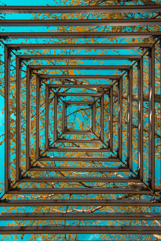 A dramatic low-angle shot looking straight up through a tall, square metal structure that creates a receding tunnel effect. Beyond the metal beams, the vibrant orange leaves of a tree in autumn are visible against a clear, bright turquoise sky.