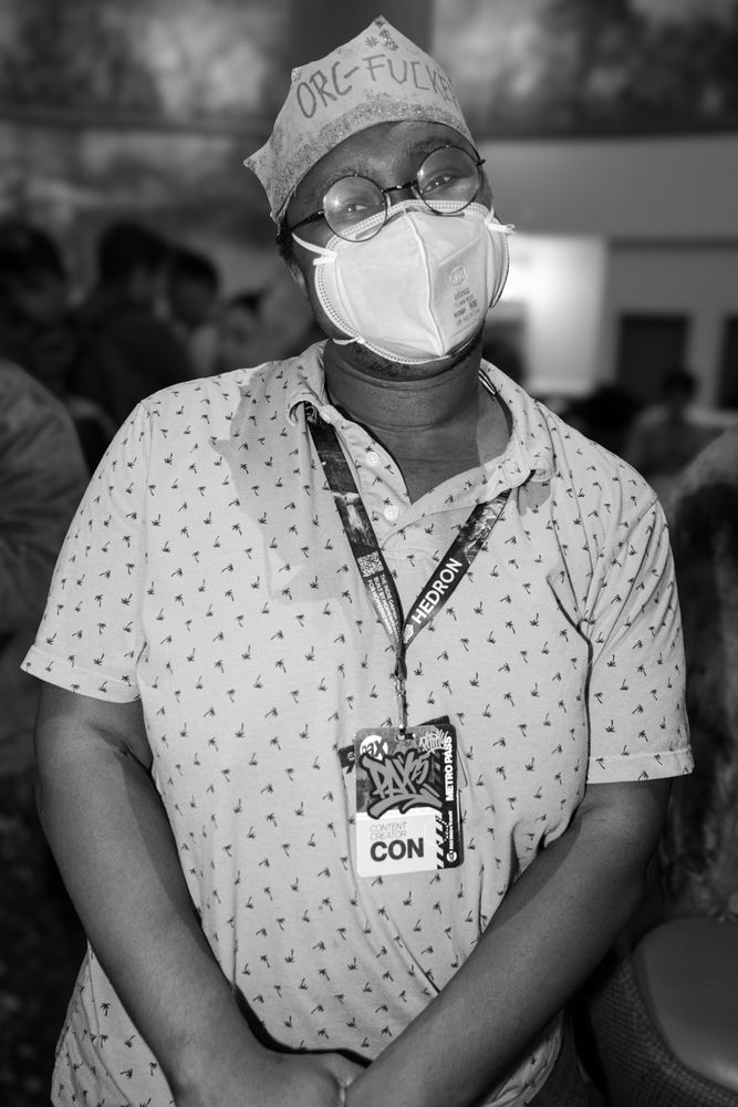 A black and white photograph of a person wearing a patterned polo shirt, glasses, and an N95 protective mask. They have a bandana or folded cloth tied on their head that reads "ORC-FUCKER". They are wearing a lanyard with an event badge and standing in a crowded indoor setting.