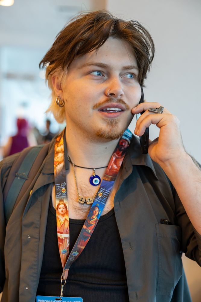 This is a candid photograph of a young person with striking light-blue eyes, captured while talking on their phone.

They have a distinctive two-toned hairstyle, with dark hair on top and bleached blonde hair underneath and at the sideburns. They are wearing a mustache and a small soul patch. Their personal style is accented with jewelry, including a silver nose ring, a hoop earring, a large, ornate silver ring on their finger, and multiple necklaces, one of which features a prominent blue and white "evil eye" pendant.

The person is dressed in a dark grey button-down shirt worn open over a black ribbed tank top. Around their neck are two colorful lanyards, similar to those seen at conventions, which appear to be for the "Gen Con" event.

The photo is a close-up, focusing on the individual's expressive face as they engage in their conversation. The background is softly blurred, indicating the photographer's focus on this single, in-the-moment slice of life.