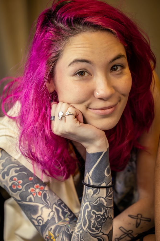 This is a close-up portrait of a woman with vibrant, hot pink hair, looking directly at the viewer with a slight smile. She has light-colored eyes and a nose piercing on her right nostril. Her left hand is propped under her chin, and she is wearing two rings on her left hand.

Both of her forearms are heavily tattooed with intricate designs in black and gray, with some hints of red and yellow on her right arm. The tattoos on her left arm appear to be a solid black sleeve with a moon and some lighter patterns emerging, while her right arm has more detailed, illustrative designs including what looks like a floral pattern. She is wearing a light-colored top, possibly beige or off-white. The background is blurred, suggesting a shallow depth of field, which keeps the focus entirely on the woman. The lighting is soft, highlighting her features and hair color.