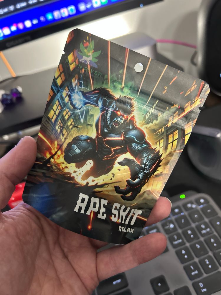 A person's hand holds a small, flexible product pouch, likely for cannabis or related products. The pouch features a graphic of an enraged, muscular ape glowing with orange-red energy lines, leaping across a ruined, night-time city backdrop. The large text on the pouch reads "APE SHIT" followed by the word "RELAX" beneath it.