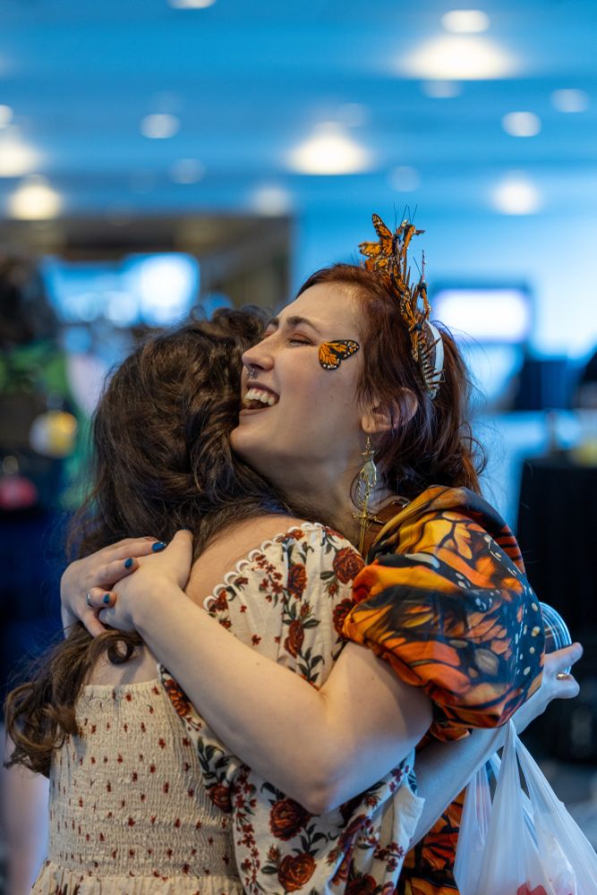 This is a heartwarming, vertical shot capturing two people in a close hug. The focus is on the person on the right, whose joyous expression is clearly visible.

This person has long reddish-brown hair styled in an intricate updo adorned with a crown or headpiece made of monarch butterflies. There's also a small butterfly decal or piece of face paint on their right cheek. They are wearing a blouse with puffy sleeves that feature a vivid monarch butterfly wing pattern. They are embracing the other person tightly, laughing with their eyes closed and their head tilted back slightly.

The person on the left, seen from behind, has long, curly dark brown hair and is wearing a light-colored, off-the-shoulder dress with a repeating floral pattern. They are also hugging the other person tightly, with their hands visible on the person's back.