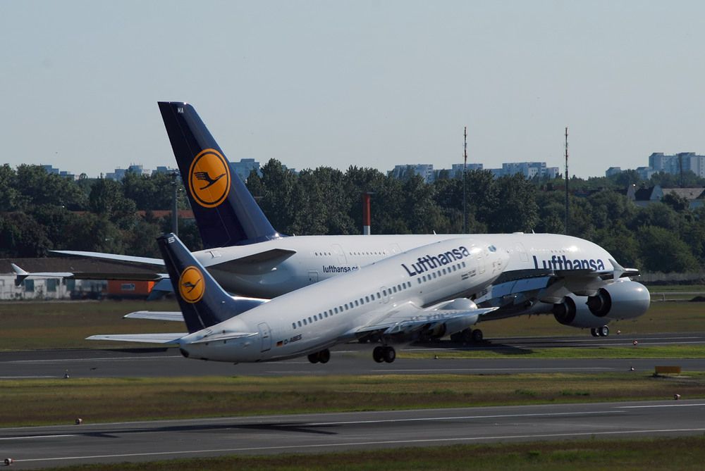 Das Bild zeigt zwei Lufthansa-Flugzeuge auf einem Flughafen. Im Vordergrund hebt ein kleineres Flugzeug ab, während im Hintergrund ein größeres Flugzeug auf der Startbahn steht. Beide Flugzeuge tragen die typische weiße Bemalung mit blauen Heckflossen und dem gelben Lufthansa-Logo. Im Hintergrund sind Bäume und einige Gebäude zu erkennen.