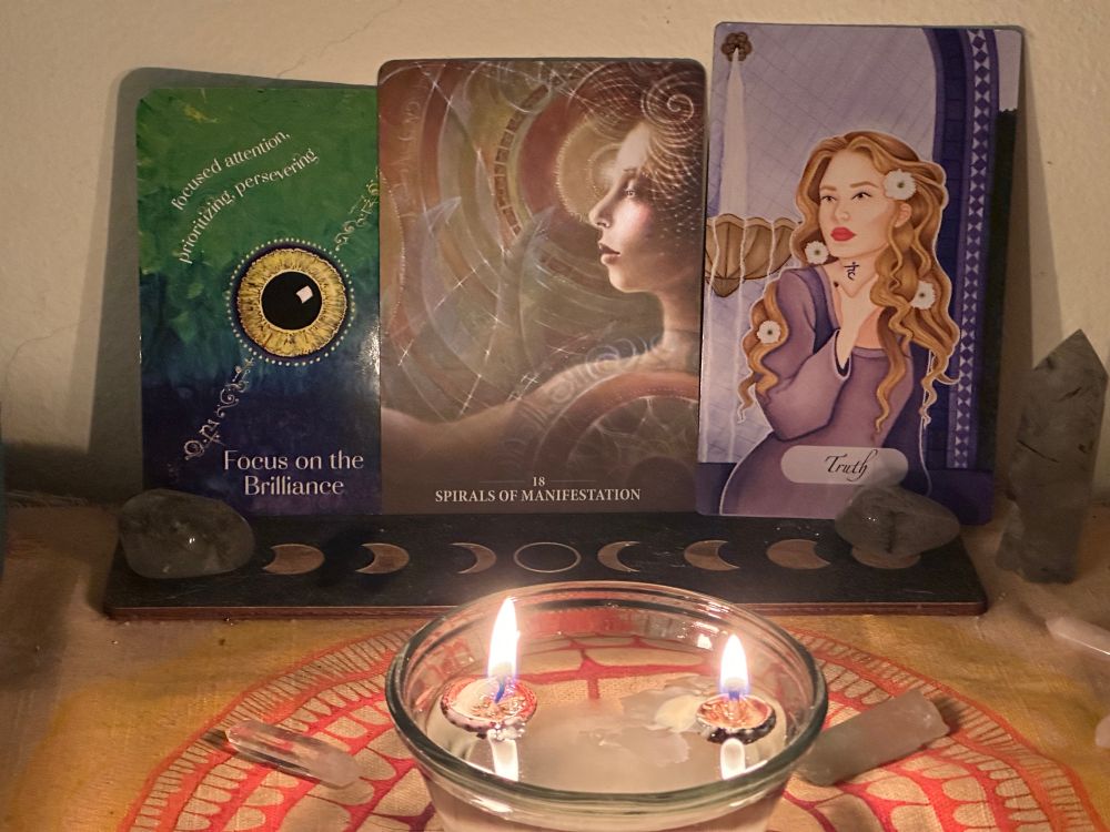 Two acorns filled with wax burn in a bowl of water. Three oracle cards stand behind. 

“Focus on the Brilliance”
“Spirals of Manifestation”
“Truth”