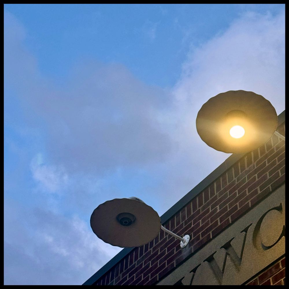 A low-angle shot of two large, cantilevered industrial barn lights on a red brick building. The building has partially obscured carved stone lettering that reads "YWCA." The light on the far right is on, casting a warm yellow glow, while the other to the left is missing its light bulb. The backdrop is an early morning sky with soft blue and wispy, side-lit clouds.