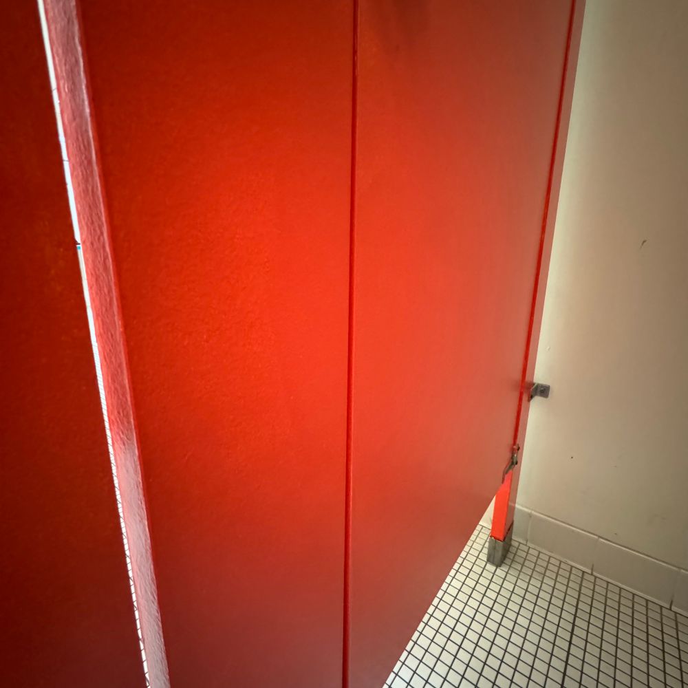 A vibrant orange restroom stall door, viewed from inside, with a white wall and a white tiled floor below.