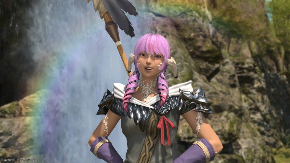 A screenshot from Final Fantasy 14. This is a screenshot of one of my newest characters, Melancholia Wyrmborn. Melancholia is a female Raen Aura with tanned skin, hot pink eyes and purple hair with pink accents. Her hairstyle has a fringe and one large swirl on each side, with a braid on the back. In the picture, Melancholia is in the Central Shroud, in front of a small waterfall, she is seen from head to waist, and there's a small rainbow behind her on the left side. She is smiling and her hands are on her hips. She is wearing the starting Au Ra top and purple forearm-lenghthed gloves. In her back she has a spear with two grey feathers on top.