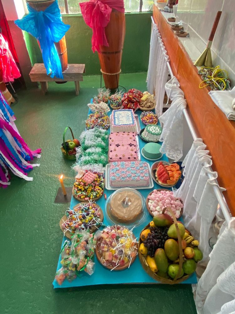 mesa no chão cheia de bolos, doces e frutas, na frente de um altar de terreiro de umbanda e ao lado de atabaques
