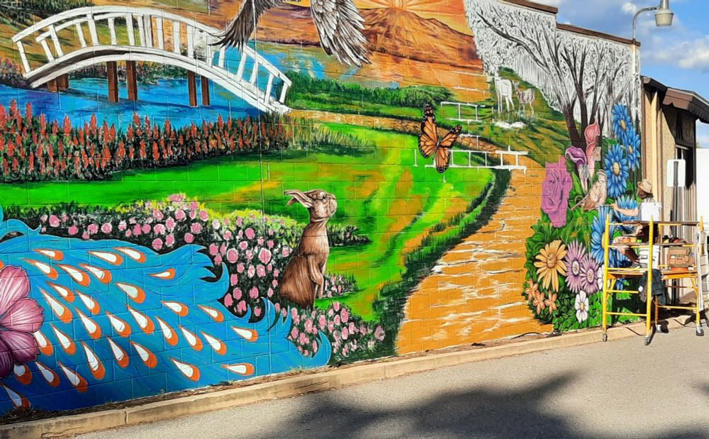 On the right, an artist on a scaffold is painting large wall mural. Flowers on the right sideli e a brick walk that curves to the upper left and a white bridge over a small stream.  A grassy area is between the walk and the stream.  Flowers line the water's  edge below the bridge and again where the stream appears in the lower left corner, where what looks like drops (maybe a small rapids?) curve downward.  An upright rabbit is in the center bottom of the scene.  The wall is painted in bright pinks, greens, yellows, oranges, and white.