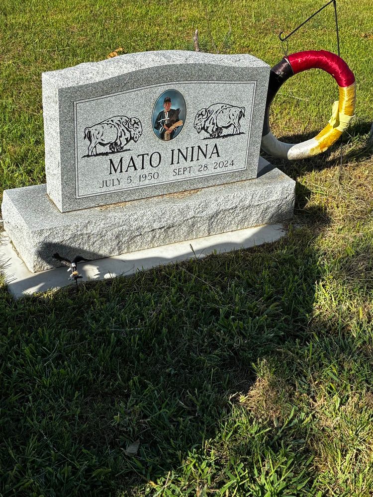 Atop green mowed grass a headstone with two buffalo in profile facing an oval photograph in the center with my uncle in his regalia and his Dakota name, MATO ININA etched into the grey granite. A medicine wheel with the four colors hangs to the right of the stone.