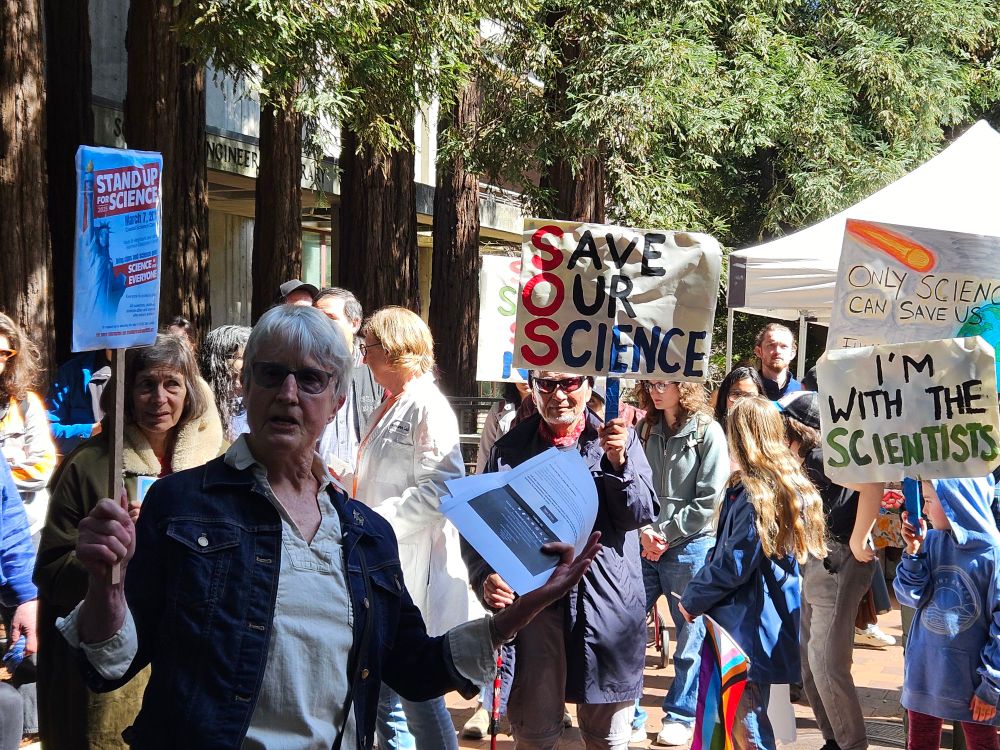 Donna Haraway, elder intellectual white woman, speaking before a tight group of people with save science signs amidst the redwoods at UCSC.