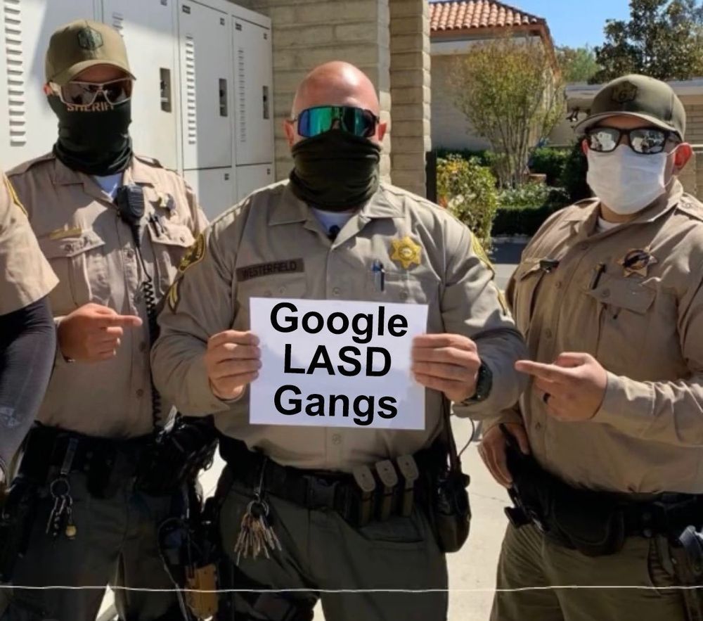 Three Los Angeles County sheriffs stand in sunglasses and masks. The two on the ends point to a piece of paper the one in the middle is holding. It reads (edited onto it) “Google LASD gangs”