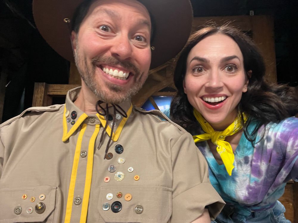 Eden and brother Sam Riegel smile and pose in a selfie. He’s wearing camping gear, she’s got a tie dye T and a yellow bandana on her neck. 