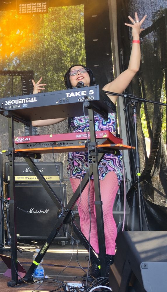 Photo of me on stage, arms up in a bhangra like dance shape behind my synth station, with my face tilted upwards, eyes closed and a happy smile. Wearing one of my fluorescent outfits and violently neon pink fishnet tights :)