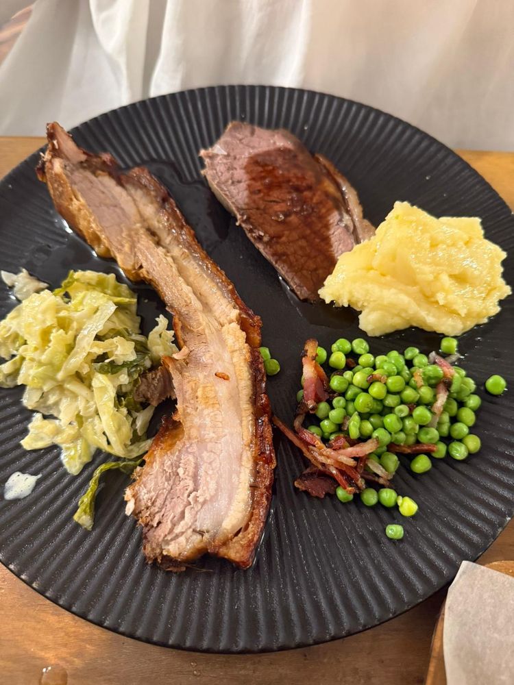 Schwarzer, geriffelter Teller mit einer Scheibe Kümmelbraten und einer Scheibe Roastbeef. Als Beilagen Kraut, Erdäpfelpüree und Erbsen mit Speck.