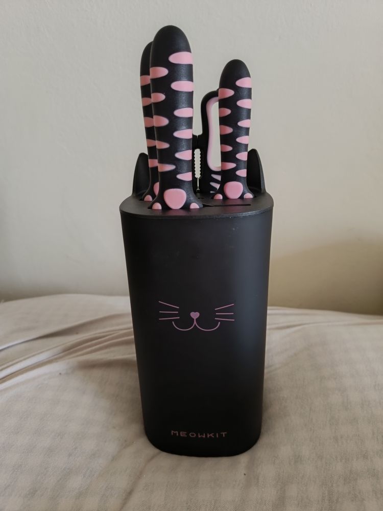 Ein schwarzer Messerblock mit einem rosa Katzengesicht und Ohren. Drinnen stecken 3 Messer und eine Schere mit schwarzen Griffen und rosa "Tigermuster" sowie Pfotenmuster am Ende des Griffes zur Klinge hin.