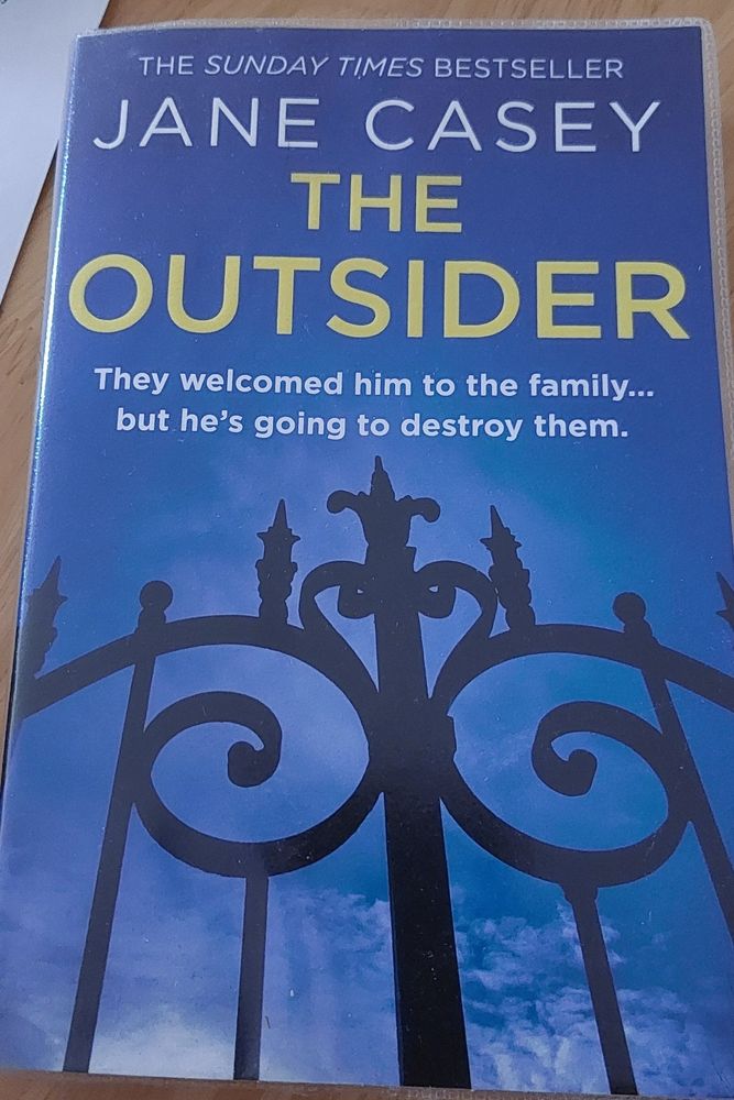 Front cover of the crime fiction book, The Outsider, written by Jane Casey. A view looking up at an ornate iron gate with a bright blue sky with a few wisps of white cloud. Yellow text for the title, The Outsider, and a sub text of 'They welcomed him into the family ... but he's going to destroy them.'