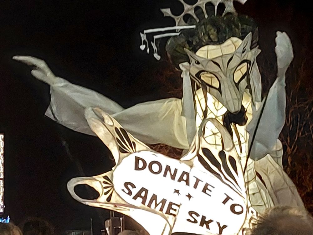 Larger than life illuminated lantern puppet in the shape of a woman, being carried above people's shoulders in a parade at night.