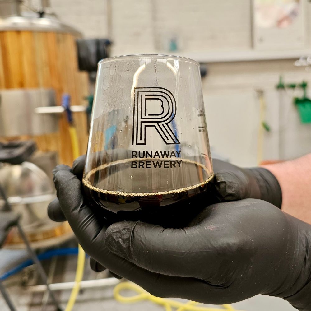 Two hands wearing black gloves are cupping a Runaway Brewery branded glass with a dark beer in it