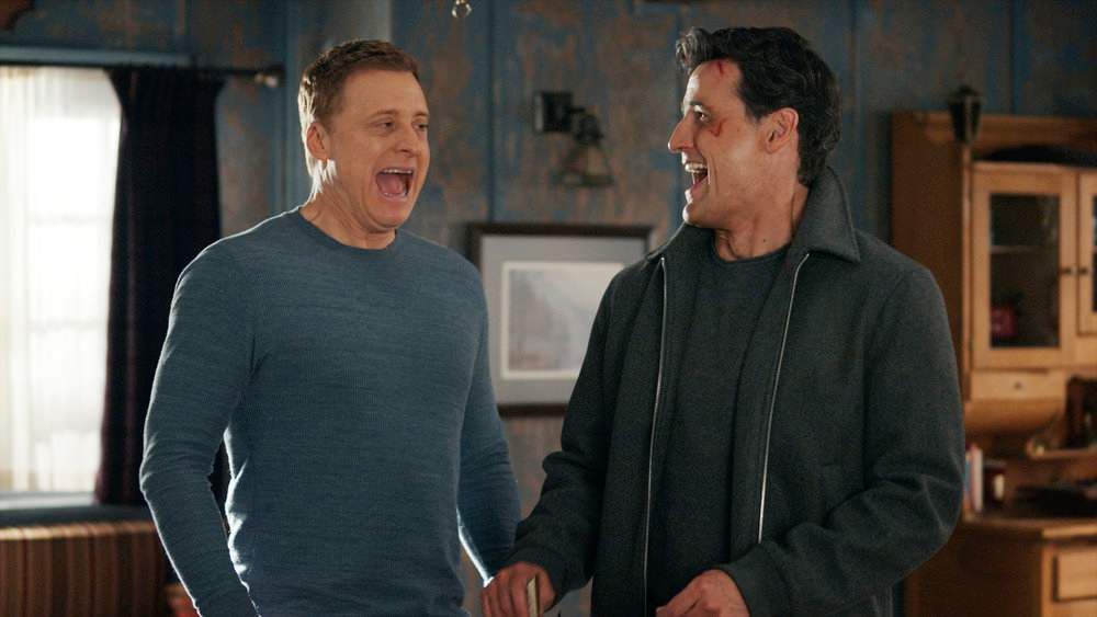 Two older men standing in a living room. The room has rusty-looking blue wallpaper and wooden decorations. One of the men has brown hair and is wearing a blue sweater, the other has black hair and is wearing a black jacket and shirt. The one with black hair has blood on his face. They are laughing.