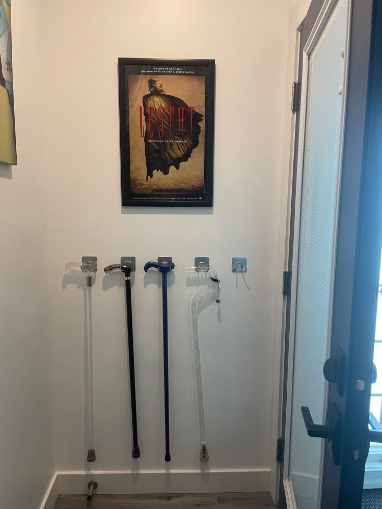 Narrow foyer with white wall. There is a Broadway poster in a black frame of "Lestat" with the tagline: "Die Young. Live Forever." Underneath are five evenly spaced hooks with four canes hanging on all but the last right hook. Left to right, the derby handle cane is clear, the next is black with a smoke derby handle, the middle is color shift blue-purple with a derby handle and the last to the right is a Neo Walk bubbles cane with a snoopy handle and charm. 
