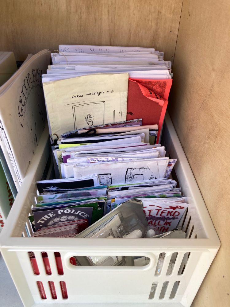photo of a white plastic bin holding zines of various sizes and colors. the bin is full of zines. there is also red cards and narcan.