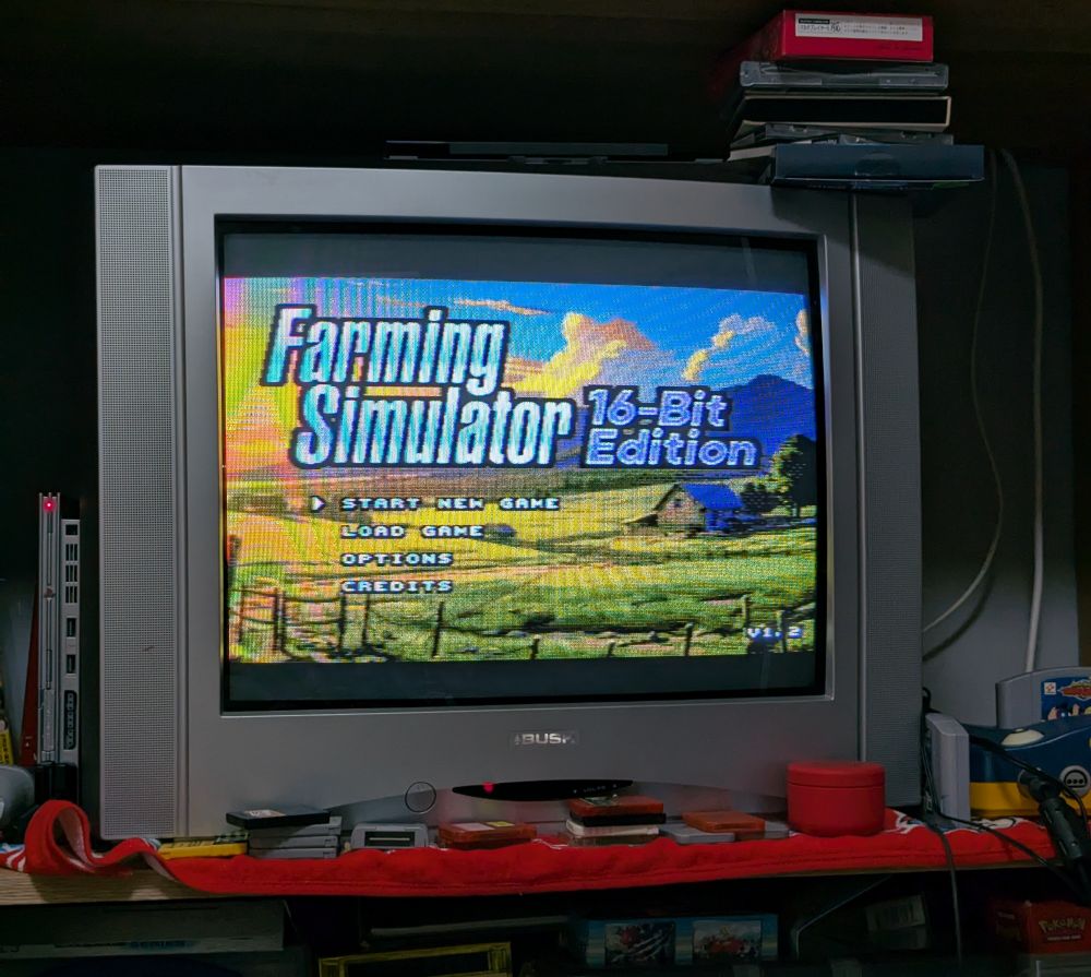 On a large CRT TV sat on a shelf surrounded by various video game paraphernalia is the start screen for Farming Simulator 16-bit Edition, a lovely pixel farm scene with the options to Start a new game, load game, options and credits. 