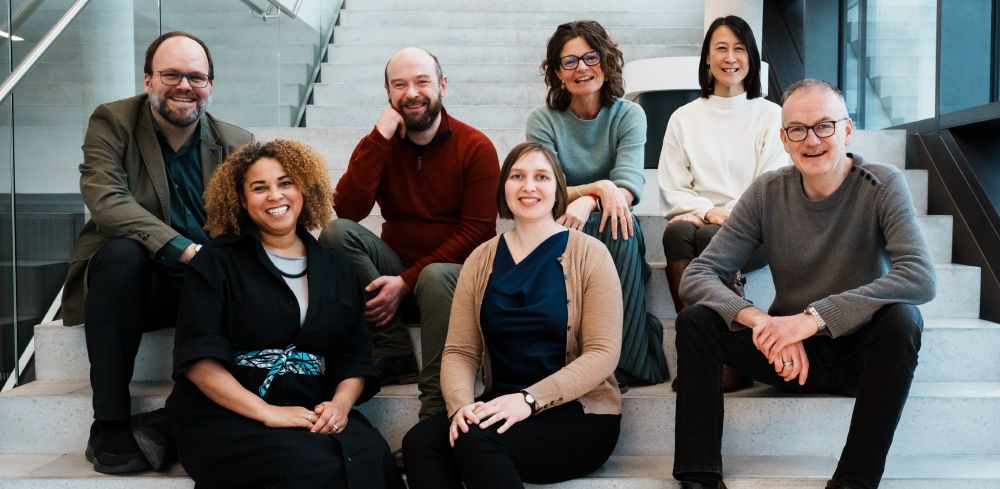 Photo of the EMBO Solutions team. The members are sitting on white steps with glass rails. From left to right: Sam, Yvonne, Aidan, Natalie, Celine, Elan, Martin. The team members are wearing smart-casual clothes and smiling, apparently at a joke that was just made.