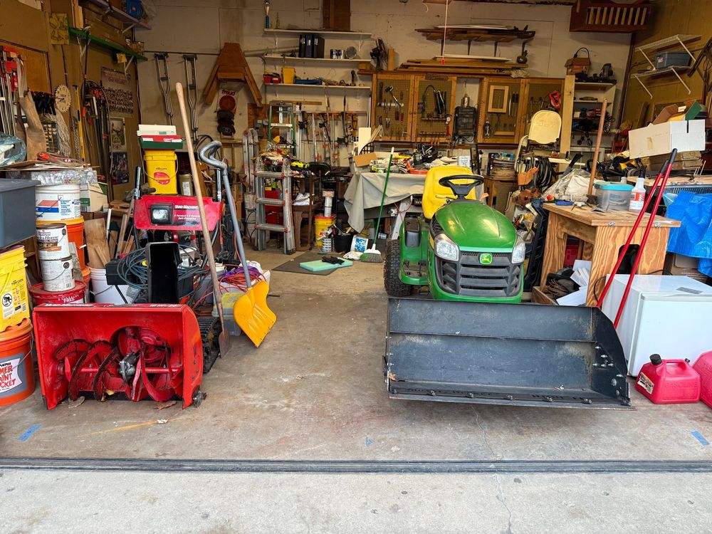 Inside of a garage with a snowthrower on the left and a lawn tractor with a bucket attachment on the right.
