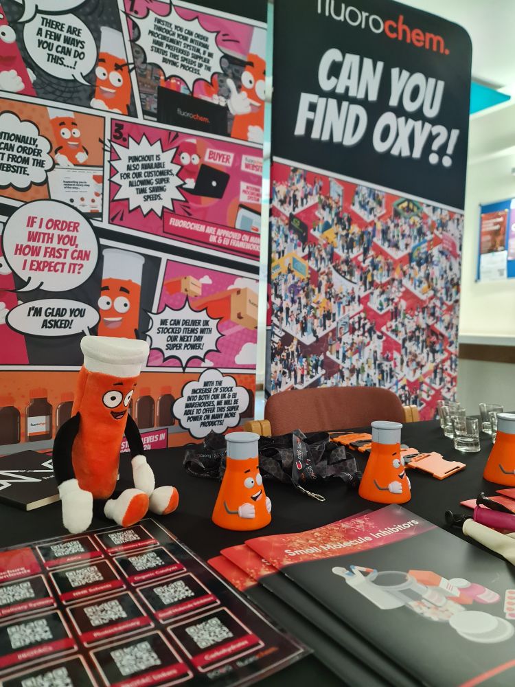 A table of orange chemistry themed merchandise including brochures about small molecule inhibitors and squishy conical flasks. The back ground is 2 banners with the Fluorochem logo and info graphics about our service