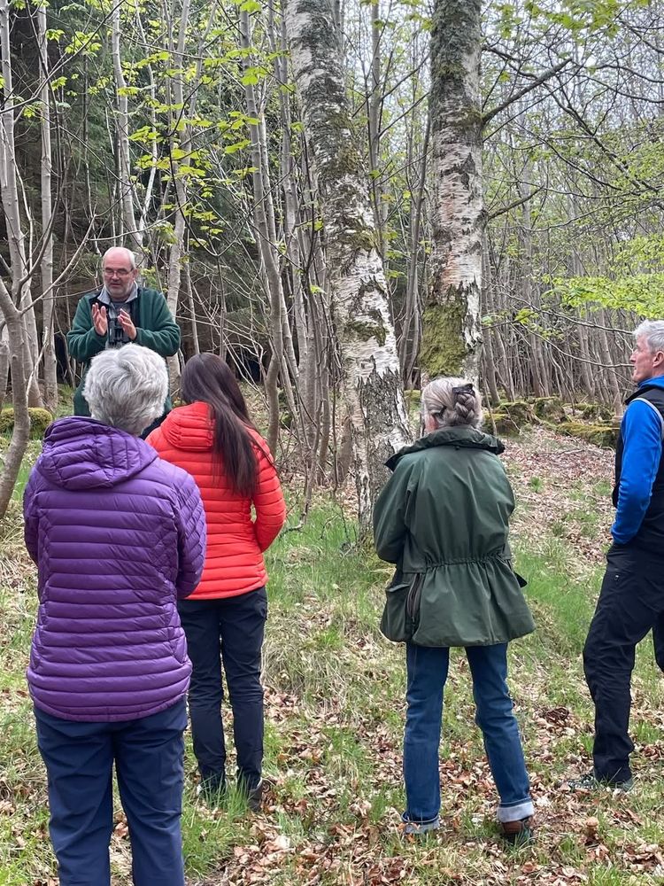 Five people stood in an open woodland setting, four facing toward trainer who is explained about camera trap workings 