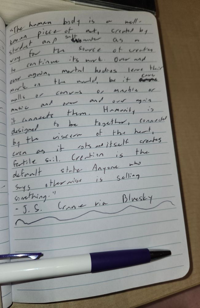 A photo of a notebook with lined paper. A purple and white ballpoint pen is laid beneath it. The quote reads "The human body is a well-known piece of art, created by stardust and salt water as a way for the source of creation to continue its work. Over and over again, mortal bodies leave their mark on the world, be it cave walls or canvass or marble or music and over and over again it connects them. Humanity is designed to be together, connected by the viscera of the heart, even as it rots and itself creates fertile soil. Creation is the default state. Anyone who says otherwise is selling something." It's is attributed to "J.S. Crane via Bluesky"
