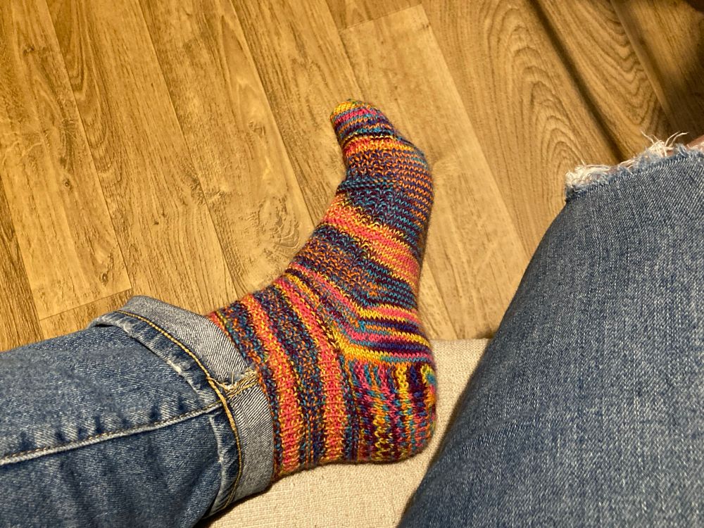 Kat’s foot wearing a multicolored textured sock. Predominant colors are pink and yellow with blue, orange, and purple also appearing. 