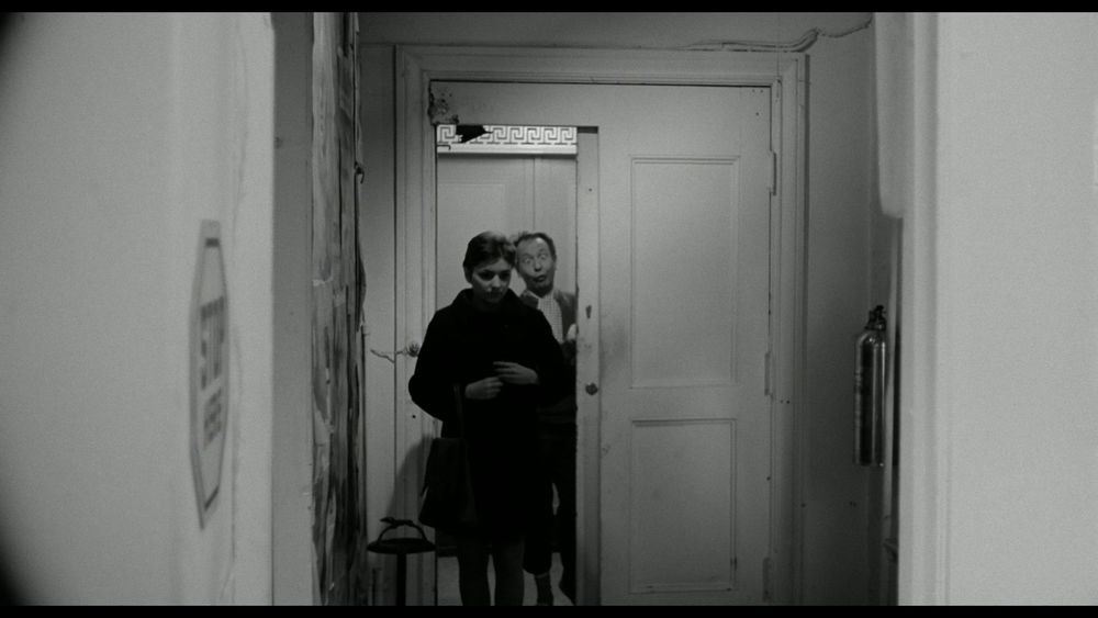 A dark-haired woman in a dark coat, framed in shadow, eyes downcast, walks away from an open elevator. Behind her, the elevator attendant is pulling an exaggerated pop-eyed leer as he stares at her butt. Off to the left on the wall is an octagonal sign that reads STOP HERE. To the woman's right is a wall-mounted fire extinguisher. 