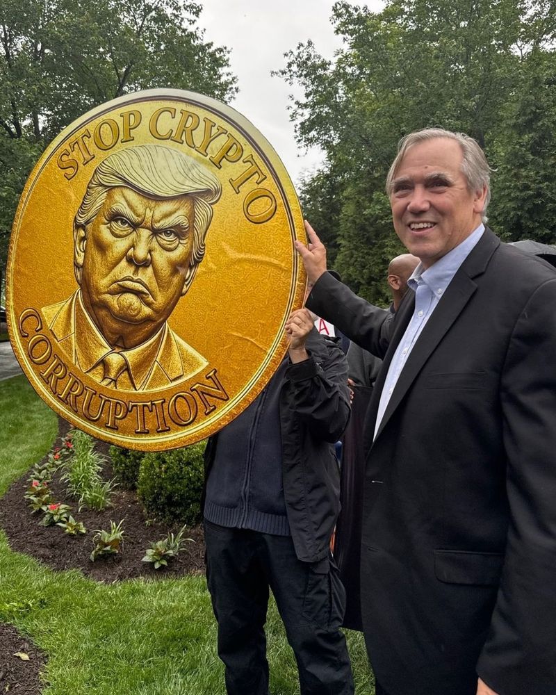 Jeff Merkley is standing next to a coin-shaped poster with an image of Trump that says Stop Crypto Corruption.