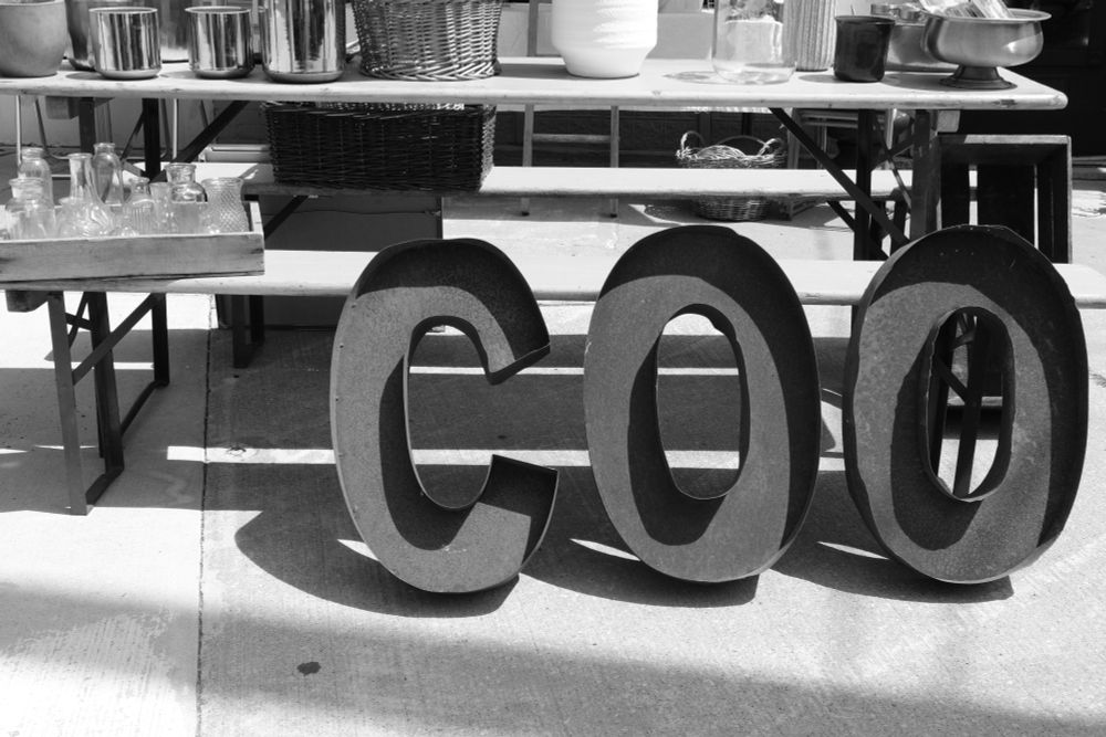 Outside a shop, various goods are displayed on tables. Sitting on the ground and leaning against a table are three large metal letters that must have been part of an outdoor sign at some point. The letters read COO.
